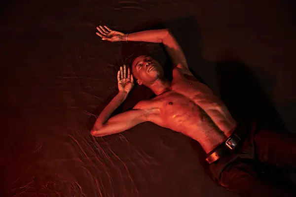 Sexy african american man lying topless on water with closed eyes, dark lights, fashion concept — стоковое фото