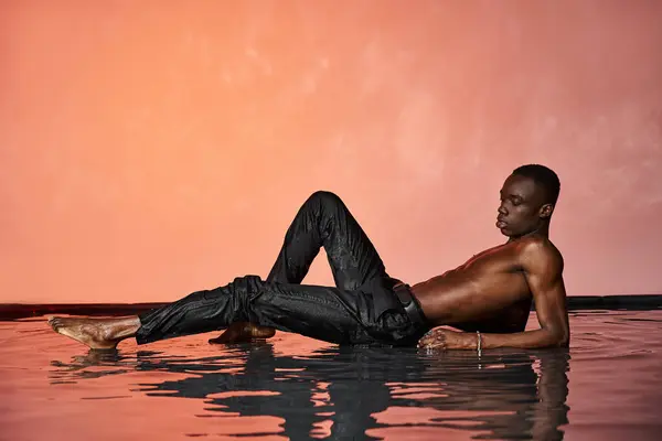 Hombre afroamericano de buen aspecto en pantalones húmedos negros reclinados en la superficie del agua y mirando hacia otro lado - foto de stock