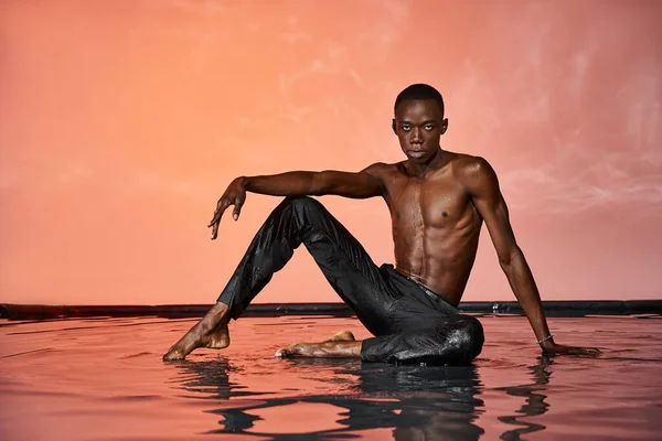 Guapo sin camisa africano americano hombre en negro mojado pantalones posando en el agua y mirando a la cámara - foto de stock