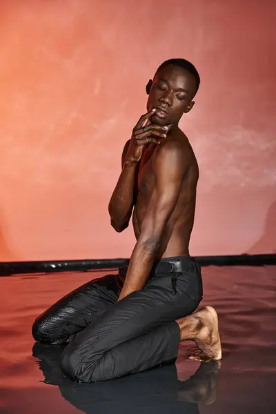 Atractivo hombre afroamericano en pantalones húmedos negros sentado en el agua rodeado de luces vívidas - foto de stock