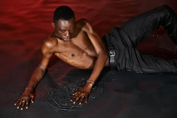 Buen aspecto sexy africano americano hombre en pantalones mojados reclinado en la superficie del agua en luces rojas - foto de stock