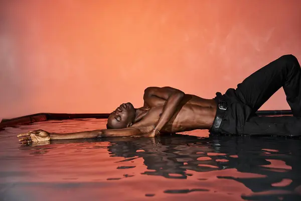 Sexy africano americano hombre en negro mojado pantalones acostado en agua superficie con cerrado ojos, rojo luces - foto de stock
