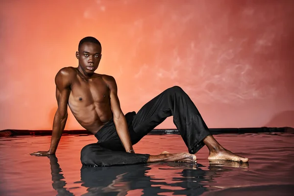 Hombre afroamericano guapo sentado en topless en la superficie del agua y mirando a la cámara, luces - foto de stock