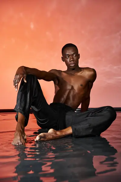 Atractivo hombre afroamericano sentado en topless en la superficie del agua y mirando hacia otro lado, luces rojas - foto de stock
