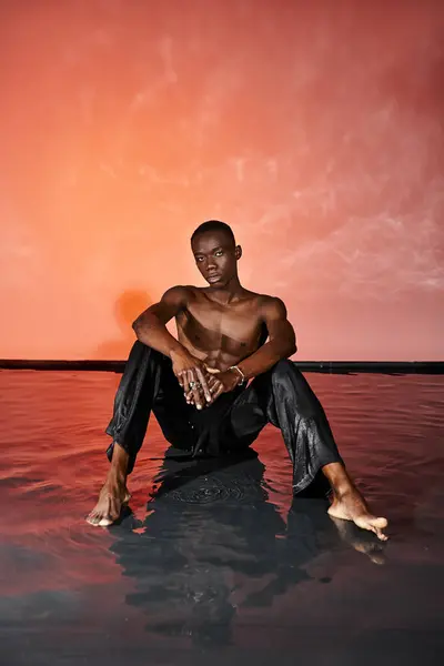 Hombre afroamericano guapo sentado en topless en la superficie del agua y mirando a la cámara, luces rojas - foto de stock