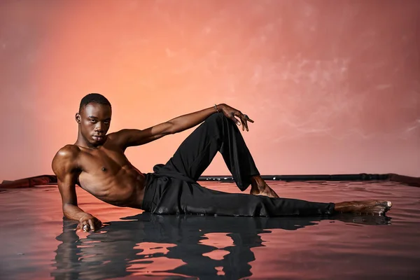 Atractivo hombre afroamericano reclinado en topless en la superficie del agua y mirando a la cámara, luces rojas - foto de stock