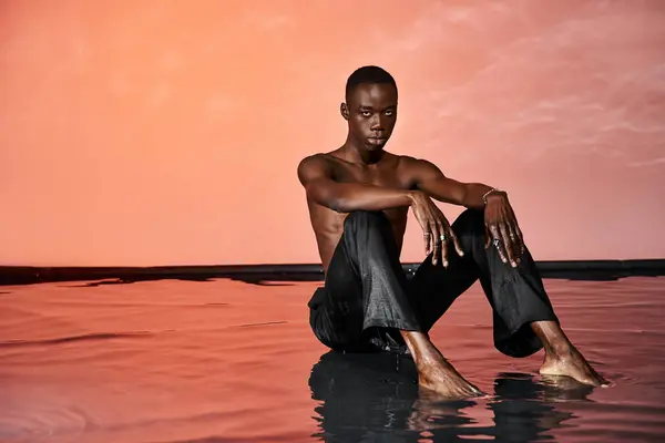 Alluring african american man in wet pants sitting on water surface and looking at camera, red light — Stock Photo