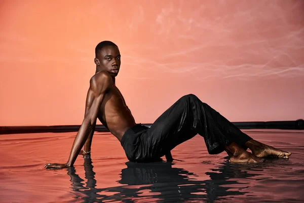 Attraente uomo africano americano sdraiato in topless sulla superficie dell'acqua e guardando la fotocamera, luci rosse — Foto stock