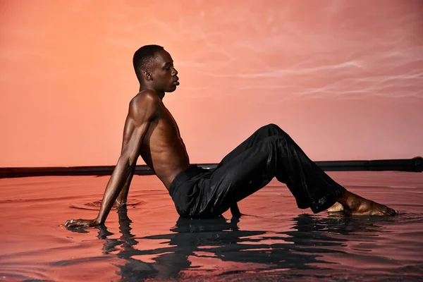 Buen aspecto sexy africano americano hombre en pantalones mojados reclinado en la superficie del agua en luces rojas - foto de stock