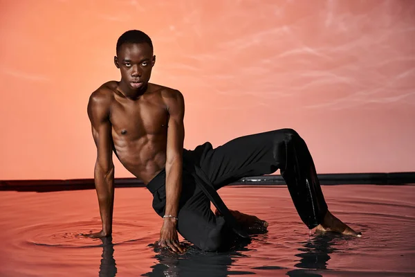 Seducente afro-americano in pantaloni bagnati seduto sulla superficie dell'acqua e guardando la fotocamera, luce rossa — Foto stock
