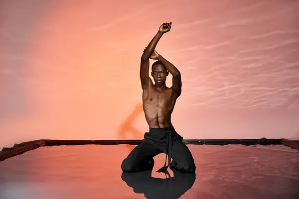 Attraente afroamericano uomo in pantaloni bagnati seduto sulla superficie dell'acqua e guardando la fotocamera, luci — Foto stock
