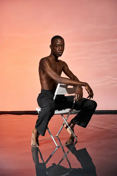 Atractivo sexy africano americano hombre sentado sin camisa en la silla y mirando a la cámara, luces rojas - foto de stock