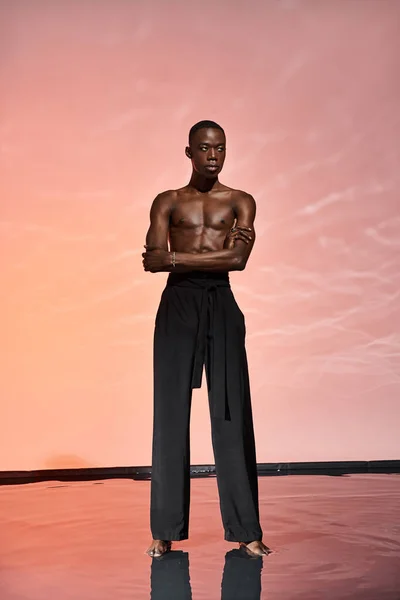 Sexy afro americano uomo in piedi senza maglietta in pantaloni neri circondato da luci e guardando altrove — Stock Photo