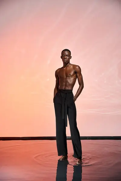 Encantador hombre afroamericano sin camisa de pie y mirando a la cámara rodeada de luces rojas - foto de stock