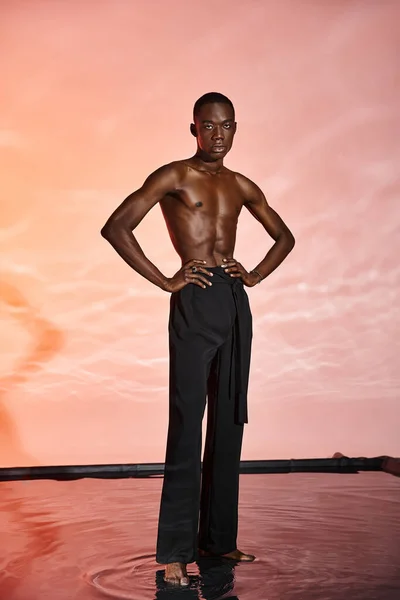 Seductor hombre afroamericano sin camisa de pie y mirando a la cámara rodeada de luces rojas - foto de stock