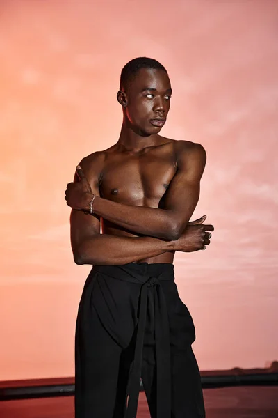 Sexy homme afro-américain debout torse nu dans un pantalon noir entouré de lumières et regardant loin — Photo de stock