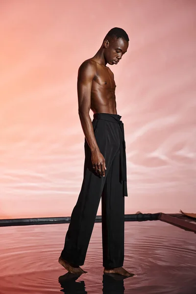 Hombre afroamericano sin camisa de buen aspecto que usa pantalones negros elegantes rodeados de luces rojas - foto de stock