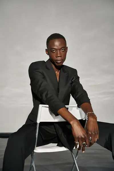 Alluring sexy african american man in elegant black suit sitting on chair and looking at camera — Stock Photo