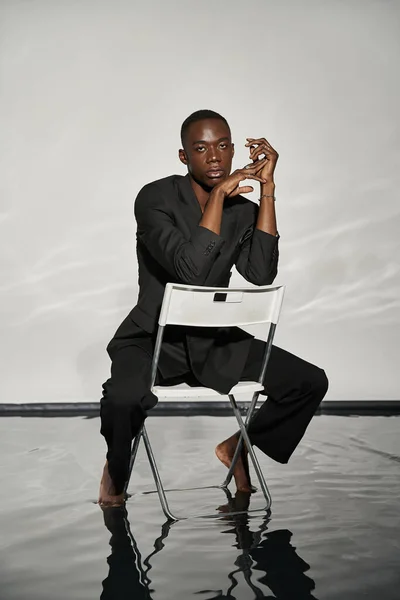 Atractivo hombre afroamericano sexy en elegante traje negro sentado en la silla y mirando a la cámara - foto de stock