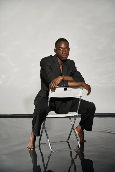 Guapo sexy afroamericano hombre en elegante traje negro sentado en la silla y mirando a la cámara — Stock Photo