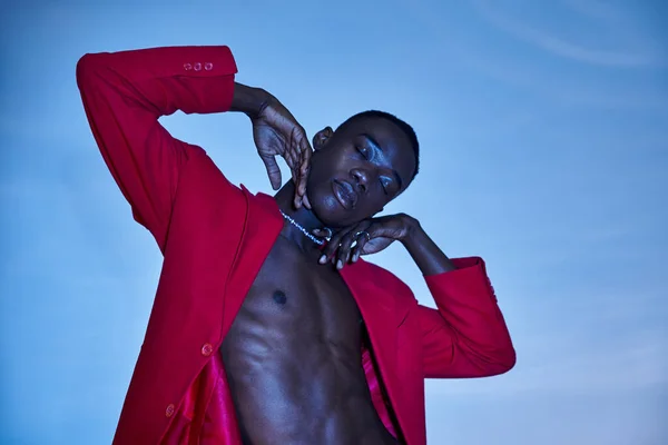 Stylish african american man in red vibrant blazer posing with closed eyes on blue backdrop — Stock Photo