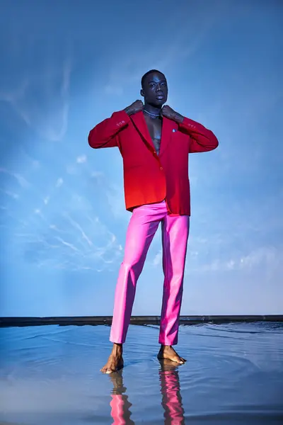 Coinvolgente elegante uomo africano americano in pantaloni rosa e blazer rosso in posa su sfondo acquoso blu — Foto stock