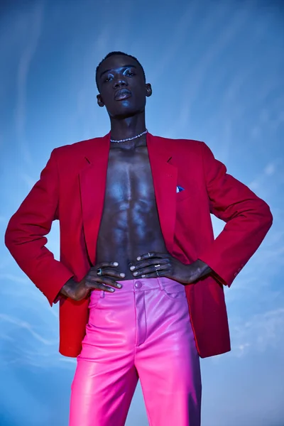 Envolvente homem americano africano elegante em calças rosa e blazer vermelho posando em pano de fundo aquoso azul — Fotografia de Stock
