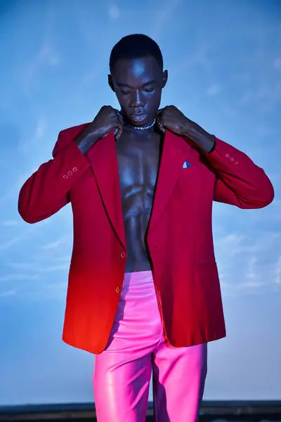 Hombre afroamericano de moda en pantalones rosas y chaqueta roja mirando hacia otro lado en el fondo acuoso - foto de stock