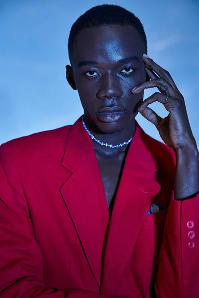 Atractivo joven afroamericano hombre en rojo vibrante chaqueta posando sobre azul acuoso fondo - foto de stock