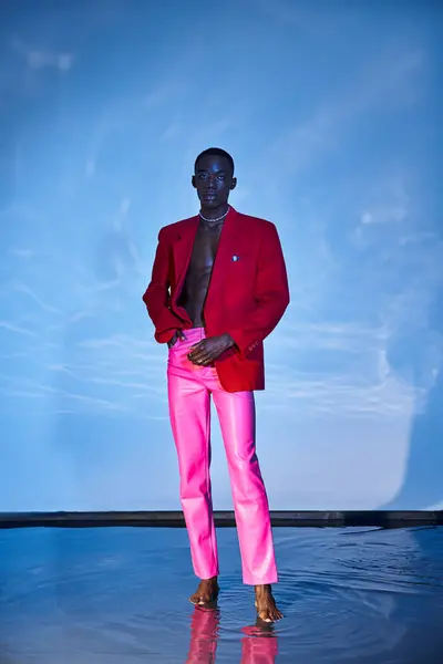 Sedutor homem americano africano elegante em calças rosa e blazer vermelho posando em pano de fundo aquoso azul — Fotografia de Stock