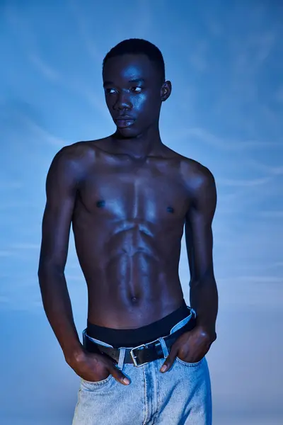Jeune homme afro-américain en jeans tendance posant sur fond bleu aqueux et détournant les yeux — Photo de stock