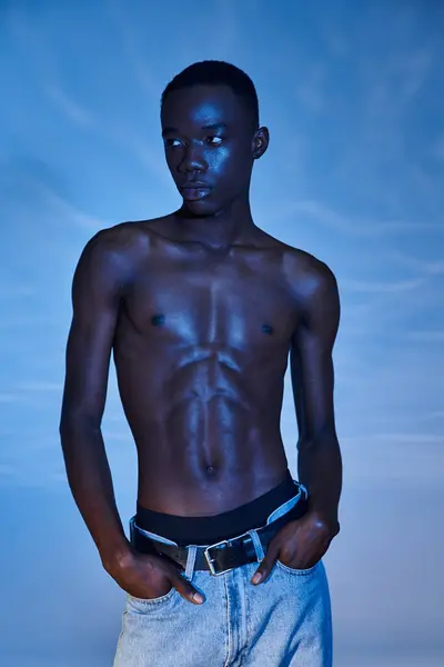 Young african american man in trendy jeans posing on blue watery backdrop and looking away — Stock Photo