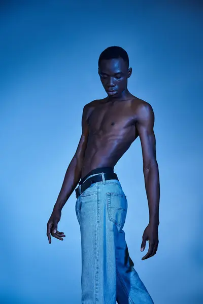 Giovane uomo africano americano in jeans alla moda in posa su sfondo acquoso blu e guardando altrove — Foto stock