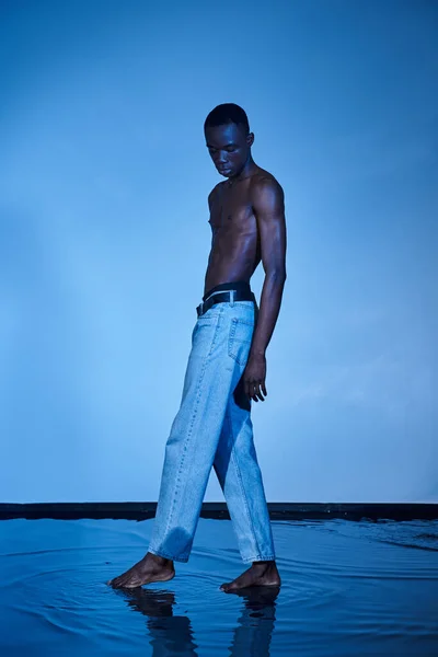 Shirtless african american man in trendy jeans posing on blue watery backdrop and looking away — Stock Photo