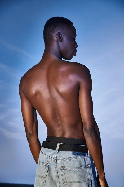 Африканский мужчина без рубашки в стильных джинсах позирует на синем водянистом фоне и смотрит в сторону — стоковое фото
