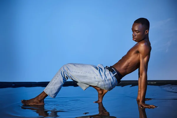 Appealing shirtless african american man in jeans posing in motion and looking away on blue backdrop — Stock Photo