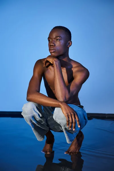 Shirtless stylish african american man in trendy jeans squatting and looking away on watery backdrop — Stock Photo