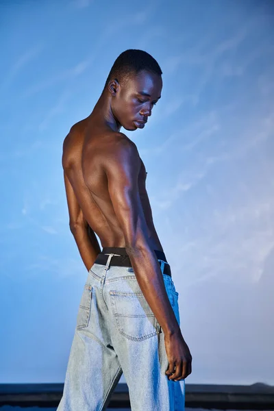Alluring young african american man in stylish jeans posing topless on watery blue background — Stock Photo