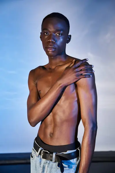 Torse nu homme afro-américain à la mode en jean élégant et en regardant la caméra sur fond bleu — Photo de stock