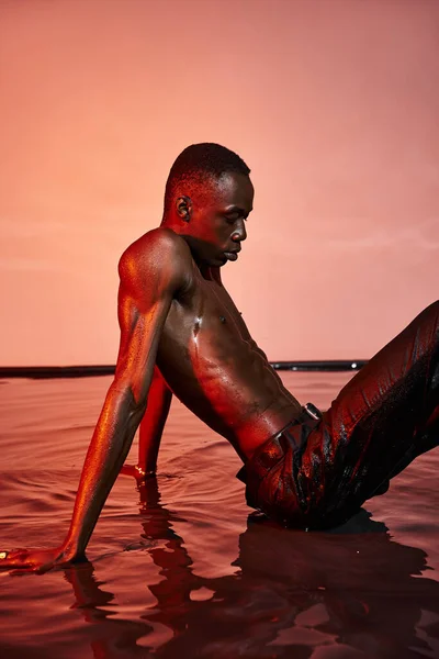 Sexy sin camisa africano americano hombre en mojado negro pantalones sentado en la superficie del agua en rojo luces - foto de stock
