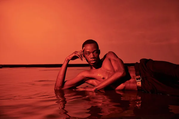 Young appealing african american man posing topless and reclining on water surface in red lights — Stock Photo
