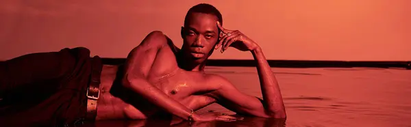 Atractivo hombre afroamericano posando en topless y reclinado en la superficie del agua en luces rojas, pancarta - foto de stock