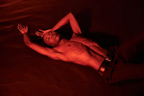 Atractivo seductor sin camisa afroamericano hombre acostado en la superficie del agua con los ojos cerrados en luces rojas - foto de stock