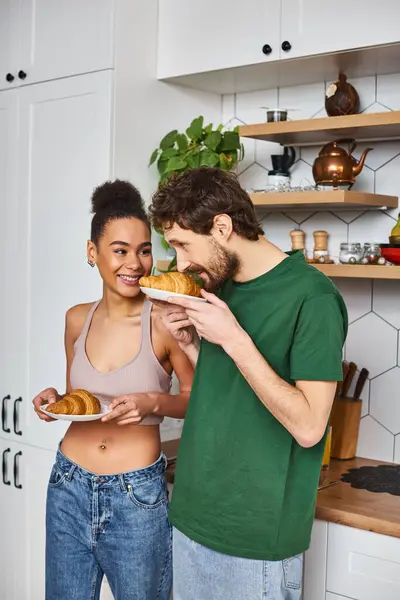 Good looking joyous diverse couple in comfy homewear holding plates with croissants in kitchen — Stock Photo