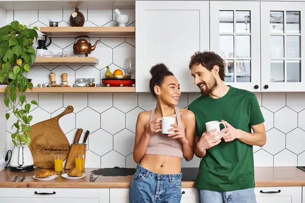 Appealing cheerful diverse couple in homewear enjoying hot coffee and looking at each other — Stock Photo