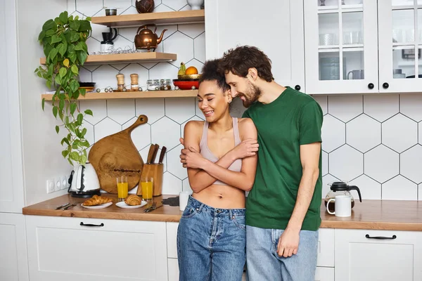 Joyous multicultural couple in homewear hugging lovingly while in kitchen before breakfast — Stock Photo