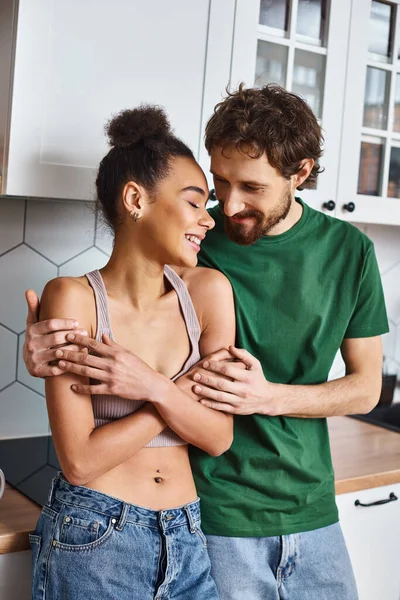 Cheerful good looking diverse couple in homewear hugging each other lovingly in kitchen at home — Stock Photo