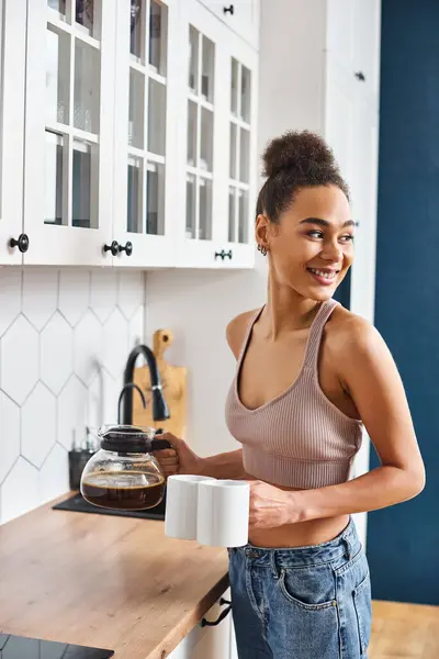 Gioiosa afro-americana bella donna in accogliente casalinghi in possesso di caffè e guardando altrove in cucina — Foto stock