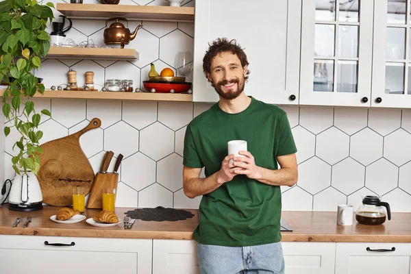 Appealing cheerful man in casual attire holding cup with hot coffee and smiling at camera joyfully — Stock Photo