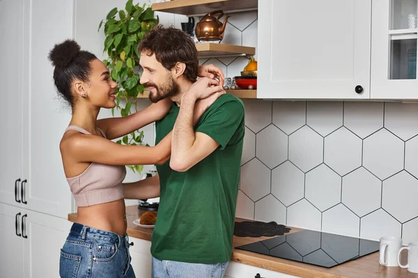 Beautiful diverse cheerful couple in cozy homewear hugging and smiling at each other in kitchen — Stock Photo